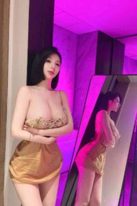 Woman in gold silk slip dress posing in front of a mirror in a purple-lit room.